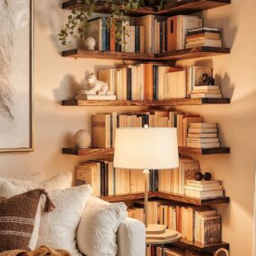 Stacked wood corner floating shelves filled with books and small objects above a cream boucle chair and reading lamp in a cozy nook.