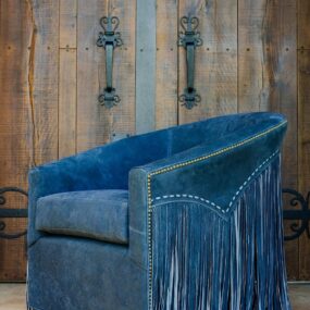 A blue suede barrel chair with long leather fringe on the arms, nailhead trim, and whipstitch detailing against a reclaimed wood plank wall.