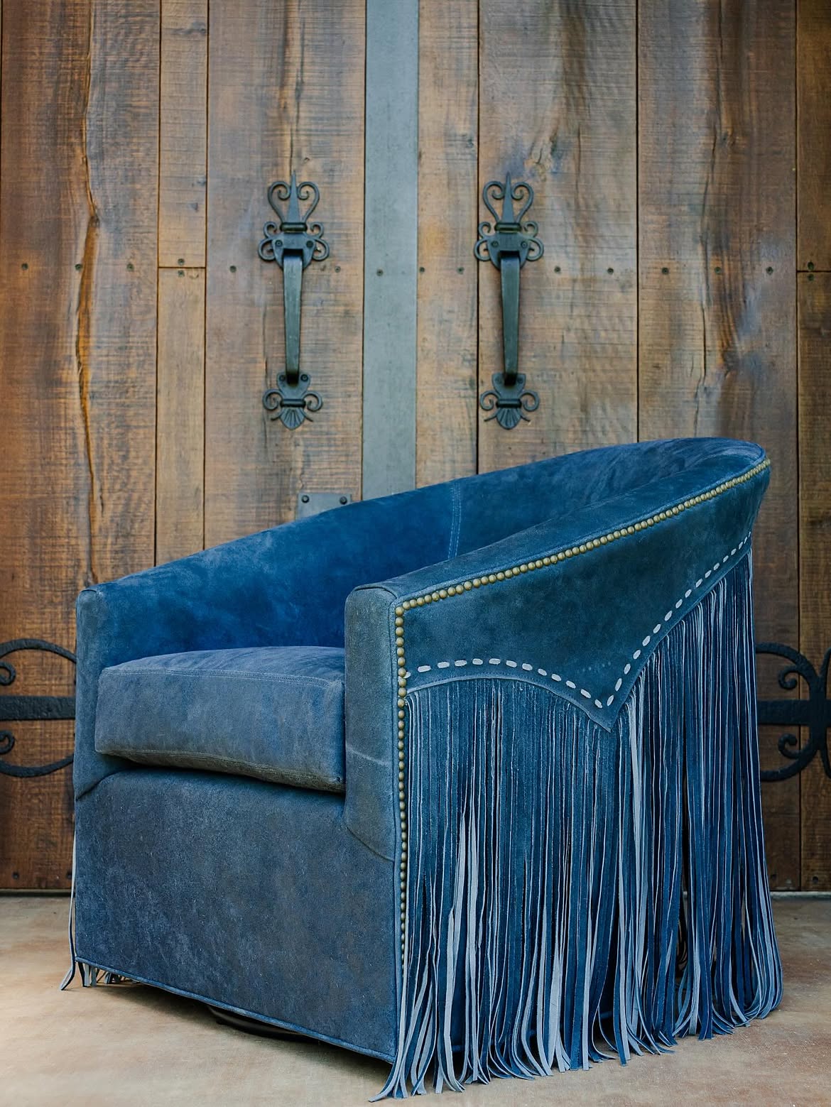 A blue suede barrel chair with long leather fringe on the arms, nailhead trim, and whipstitch detailing against a reclaimed wood plank wall.