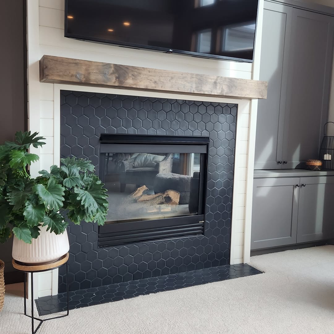 A fireplace surround with matte black hexagon tile, a chunky rustic wood mantel, mounted flat screen TV above, gray built-in cabinets to the side, and a potted plant on a stand.