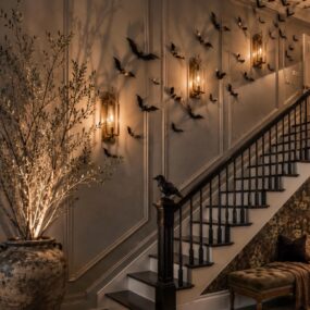 An elegant staircase wall covered in flying black paper bats, lit brass lantern sconces, a black crow perched on the newel post, a lit branch arrangement, and a tufted velvet bench.