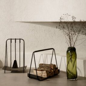 A minimalist black metal fireplace tool stand and matching log carrier holding stacked firewood, next to a tall green crinkled glass vase with dried branches against a textured plaster wall.