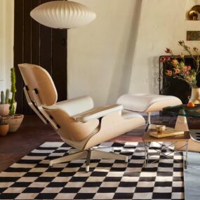 A light wood and white leather lounge chair with matching ottoman on a black and white checkered rug, under a white saucer pendant lamp in a sunlit plaster-walled room.