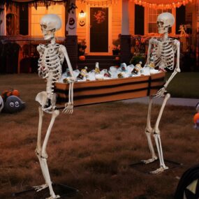 Two life-size posable skeletons carrying a coffin-shaped cooler filled with ice and drink bottles on a front lawn decorated for Halloween with pumpkins and a glowing porch behind.