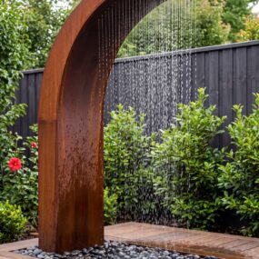 A curved rusted corten steel outdoor rain shower pouring water onto a bed of smooth black pebbles set into a wooden deck, with green shrubs and a dark fence behind.