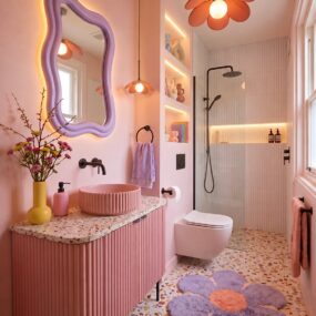 A playful pink bathroom with a fluted vanity and pink vessel sink, wavy lilac mirror, flower pendant light, terrazzo floor, and purple daisy-shaped rug.