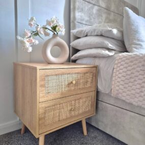 A light wood two-drawer nightstand with woven rattan drawer fronts and tapered legs, topped with a cream donut vase of pink blossoms beside a bed.