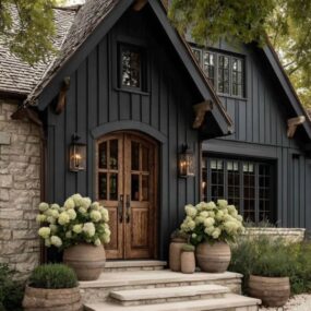 A dark board-and-batten cottage exterior with a steep gable, arched double wood front door, lantern sconces, and hydrangea-filled clay planters.