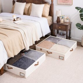 Two cream fabric under-bed storage bins with leather handles and clear tops, filled with folded sweaters beside a wood bed and nightstand.