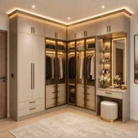 A cream L-shaped corner wardrobe with lit glass doors, a built-in vanity with backlit mirror, and a tufted stool in a walk-in closet.