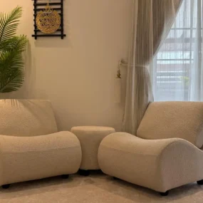 Two curved cream boucle lounge chairs with a small round ottoman between them in a sunlit sitting corner by sheer curtains and a palm plant.