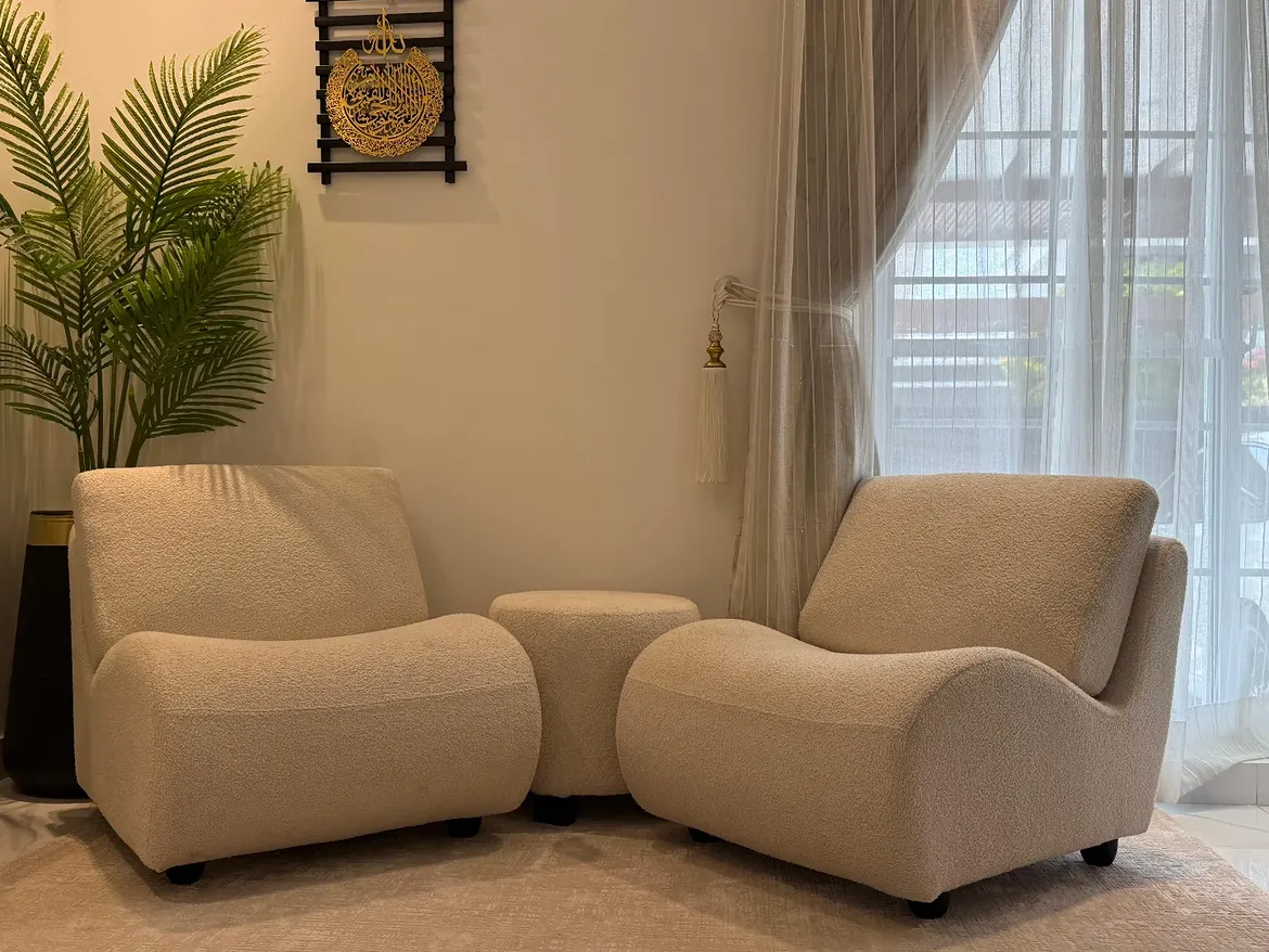 Two curved cream boucle lounge chairs with a small round ottoman between them in a sunlit sitting corner by sheer curtains and a palm plant.