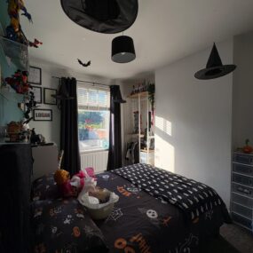 A kids bedroom decorated for Halloween with ghost and pumpkin print bedding, black witch hats hanging from the ceiling, a bat cutout, dark curtains, and a Logan name sign on the wall.