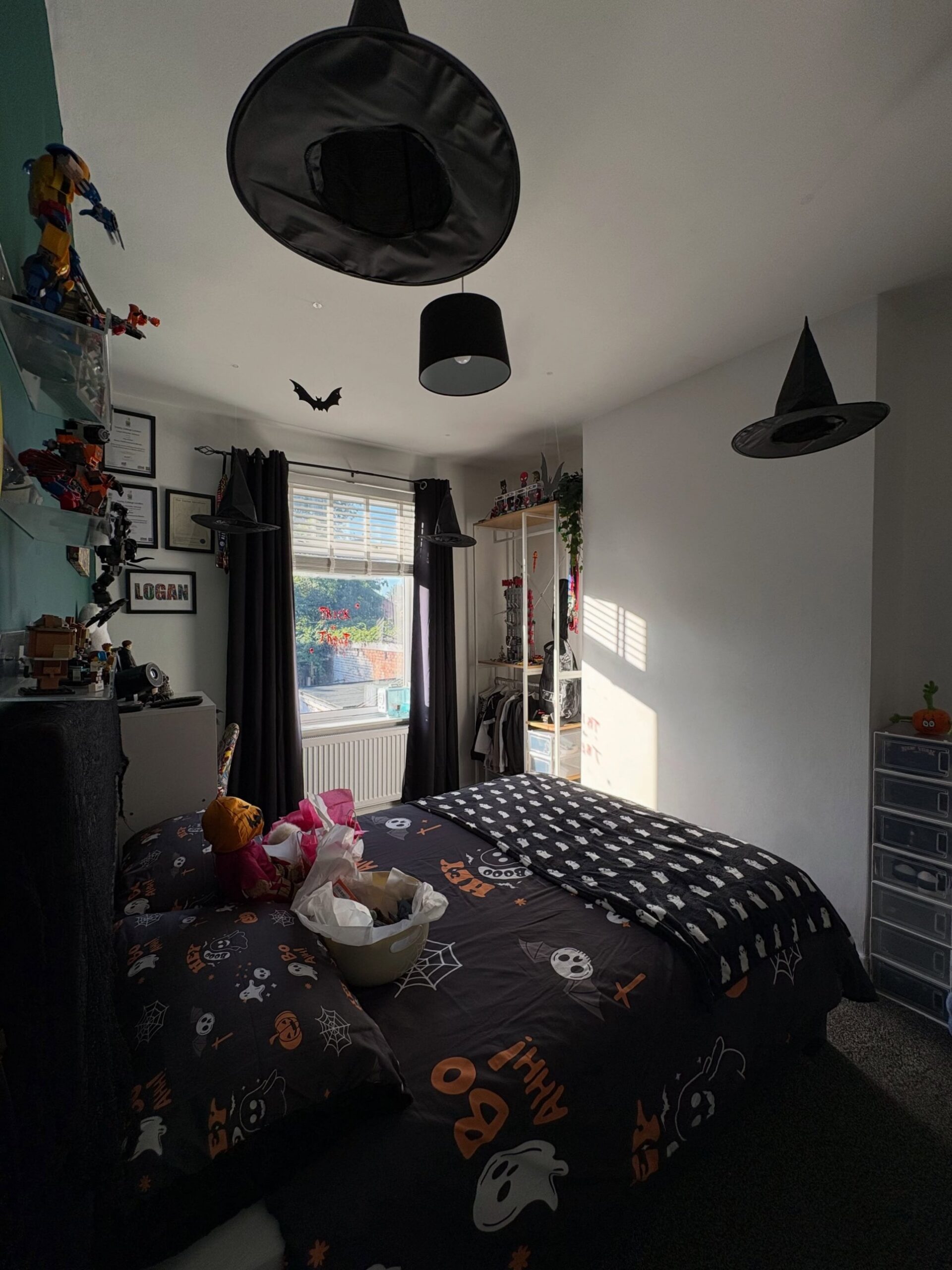 A kids bedroom decorated for Halloween with ghost and pumpkin print bedding, black witch hats hanging from the ceiling, a bat cutout, dark curtains, and a Logan name sign on the wall.