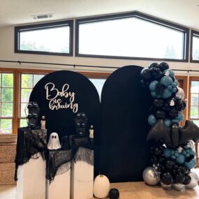 : A Halloween “Baby is Brewing” baby shower backdrop with black arches, coffin-shaped treat shelves on draped pedestals, ghost figure, pumpkins, and a black and blue balloon garland with bat and ghost accents.