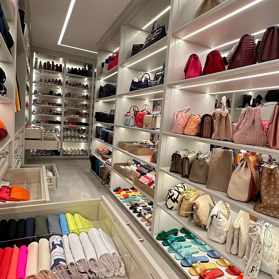 A large luxury walk-in closet with lit shelves of designer handbags, color-organized sandals, pull-out shoe drawers, and folded clothes in glass drawers.