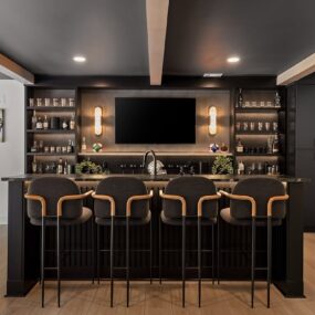 A black basement bar with four upholstered stools, a mounted TV flanked by liquor shelves and sconces, and a sports gallery wall.