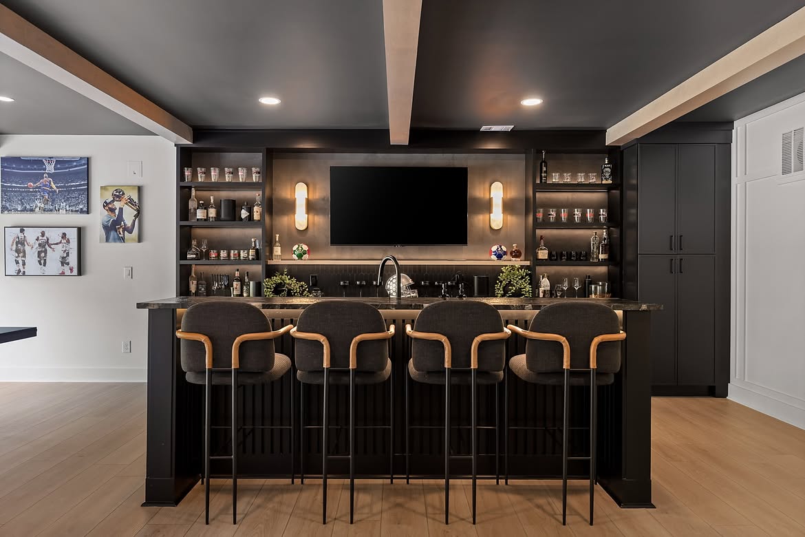 A black basement bar with four upholstered stools, a mounted TV flanked by liquor shelves and sconces, and a sports gallery wall.