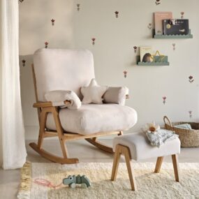 A cream upholstered rocking chair with matching ottoman beside a wall of small floral decals and sage book ledges.