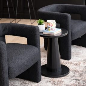 Two black boucle sculptural accent chairs with open backs flanking a round black pedestal side table on a distressed area rug.