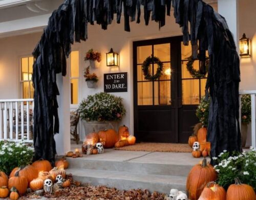 A white front porch decorated for Halloween with a tattered black cloth arch, orange pumpkins, white skulls, scattered fall leaves, and an Enter If You Dare sign by the black door.