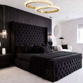 A black tufted bed with a tall wingback headboard against a black slat wall, under a double gold ring chandelier.