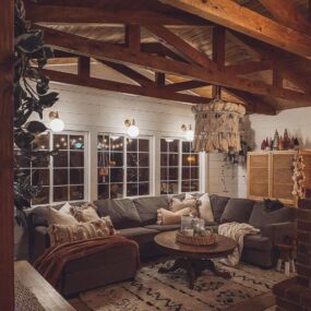 A cozy boho cabin living room at night with exposed wood ceiling beams, gray sectional sofa, macrame tassel chandelier, Moroccan rug, and tall white-framed windows.
