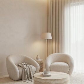 Two curved cream boucle armchairs around a round travertine drum coffee table in a soft neutral sitting corner with sheer curtains and a floor lamp.