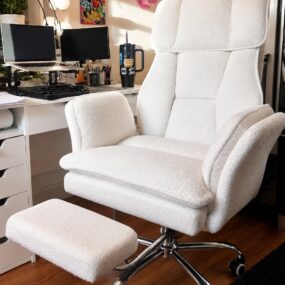 A plush white boucle reclining office chair with a pull-out footrest and chrome swivel base at a white desk with dual monitors.