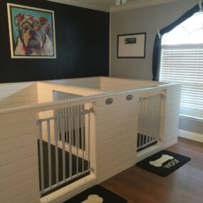 A built-in white double dog kennel with two gated doors and name plates, black woof-print mats on wood floor, and a colorful bulldog painting on a navy wall.