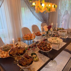 A long dining table set as a dinner party buffet with chicken wings, rice, sliders, fried snacks, dips, lit taper candles, and fresh roses under a warm chandelier.