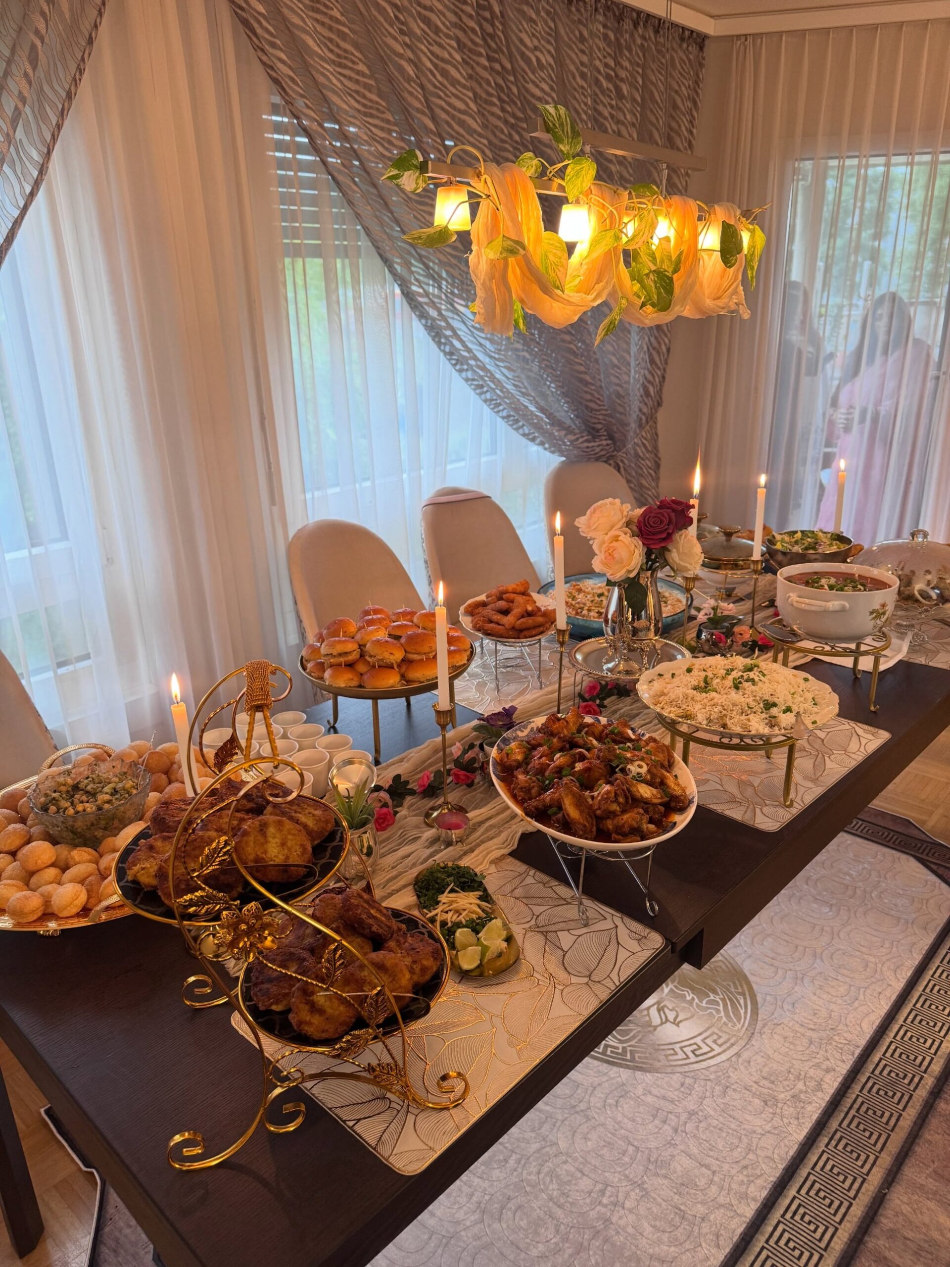 A long dining table set as a dinner party buffet with chicken wings, rice, sliders, fried snacks, dips, lit taper candles, and fresh roses under a warm chandelier.