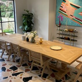 A light wood parquet dining table with cane-back chairs over a colorful chunky terrazzo floor, with abstract art and a brass bar cart behind.