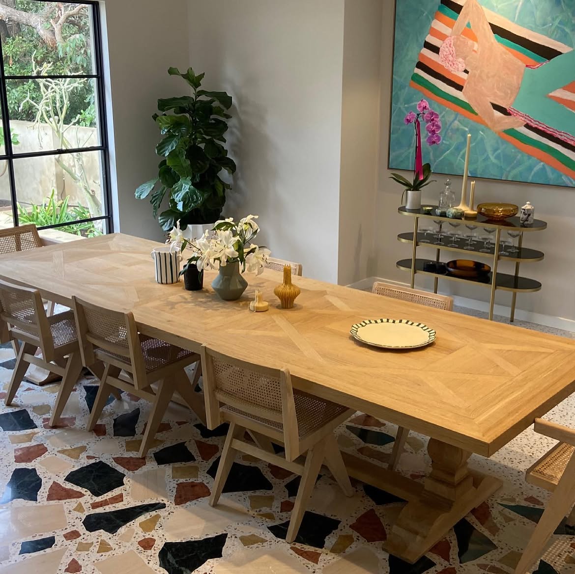 A light wood parquet dining table with cane-back chairs over a colorful chunky terrazzo floor, with abstract art and a brass bar cart behind.