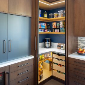 A hidden corner pantry behind dark wood cabinet doors with navy walls, wood floating shelves, LED strip lighting, and pull-out drawers of snacks.