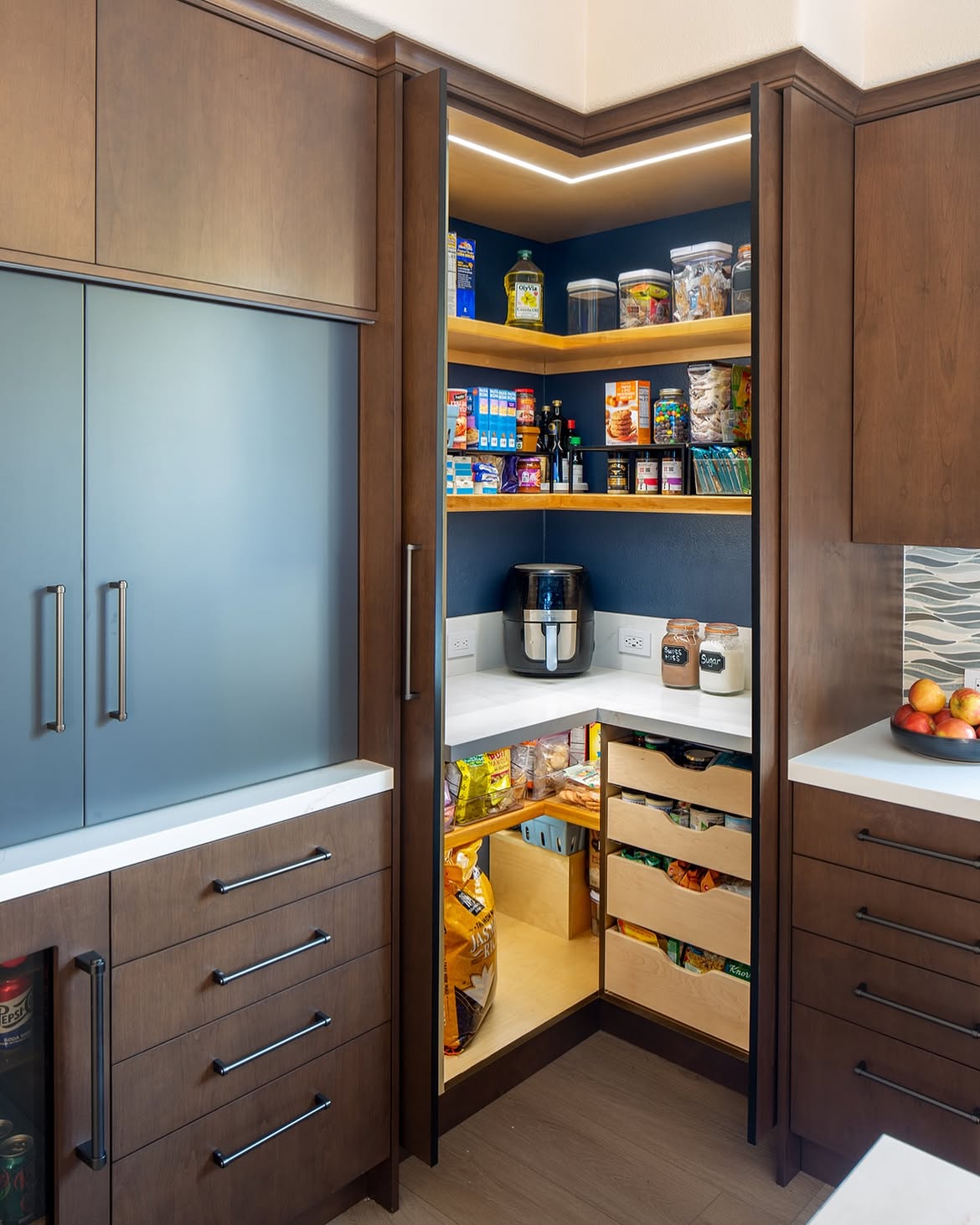 A hidden corner pantry behind dark wood cabinet doors with navy walls, wood floating shelves, LED strip lighting, and pull-out drawers of snacks.