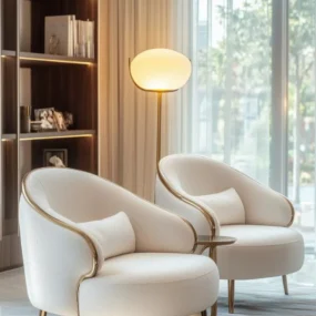 Two cream curved barrel chairs with gold trim and legs, grouped around a small side table and pebble-shaped floor lamp in a sunlit sitting area.