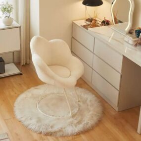 A white boucle petal-back vanity chair with gold legs sitting on a round fluffy faux fur rug beside a white dresser with a lit vanity mirror.