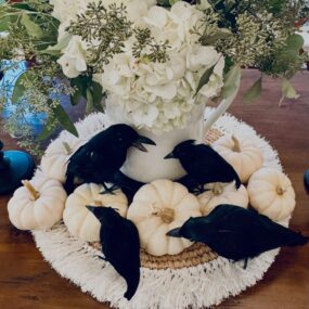 A Halloween centerpiece on a wooden table with a white pitcher of hydrangeas and eucalyptus, surrounded by white mini pumpkins and four black crow figurines on a fringed round placemat.