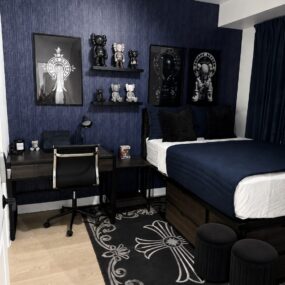 A navy and black boys’ bedroom with grasscloth accent wall, KAWS figure shelves, dark wood desk, platform bed, and black cross-print rug.