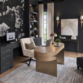 A moody home office with a black-and-white dandelion mural wall, oval wood sculptural desk, cream swivel chair, built-in bookshelves, and brass dome lamp.