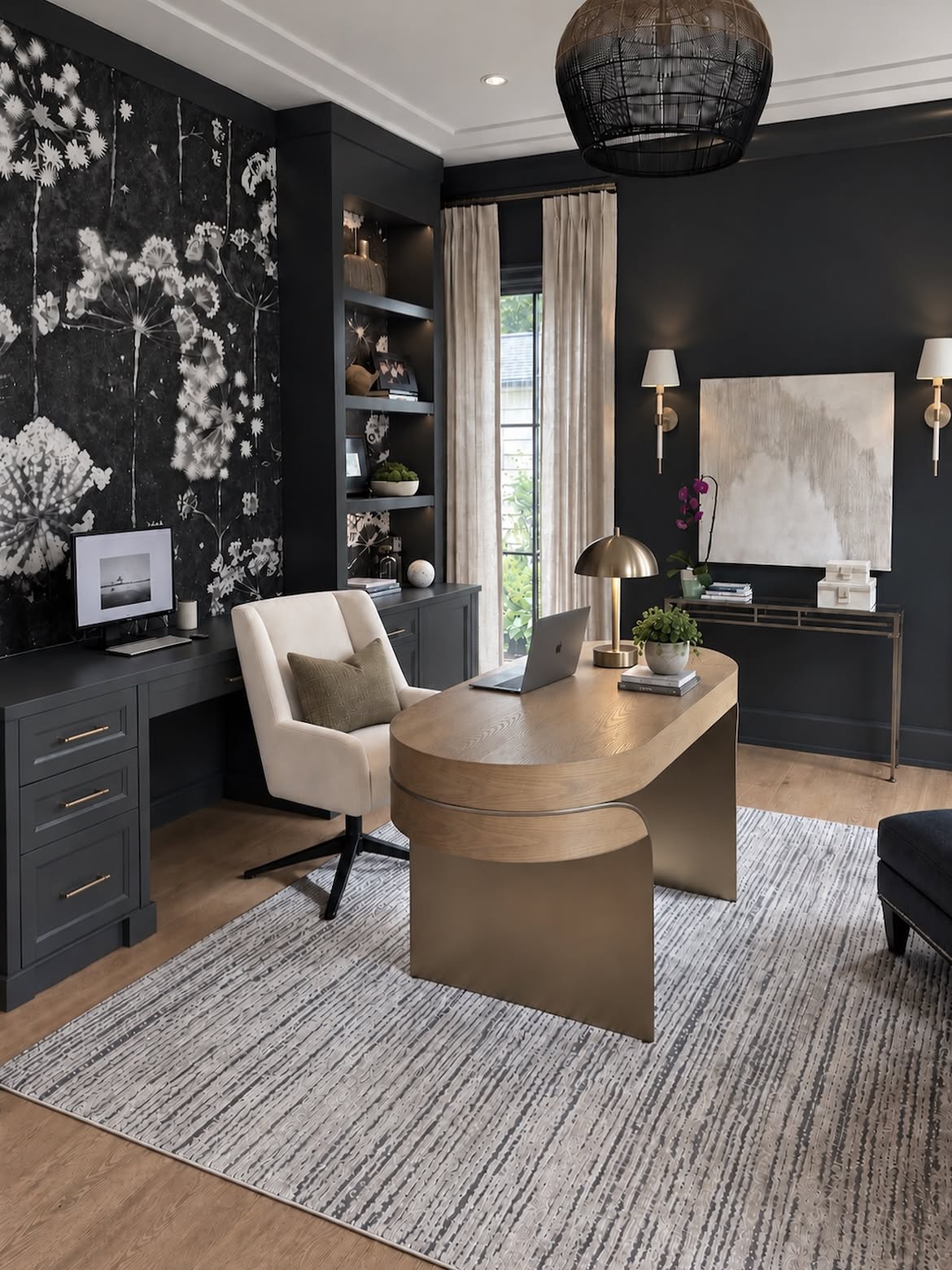 A moody home office with a black-and-white dandelion mural wall, oval wood sculptural desk, cream swivel chair, built-in bookshelves, and brass dome lamp.