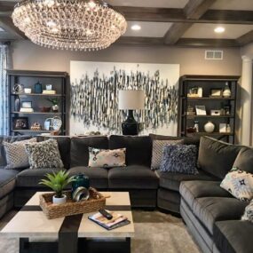 A transitional living room with a dark gray sectional sofa, round crystal chandelier, coffered ceiling with wood beams, abstract blue and gray artwork, two black bookshelves, and a two-tone coffee table.