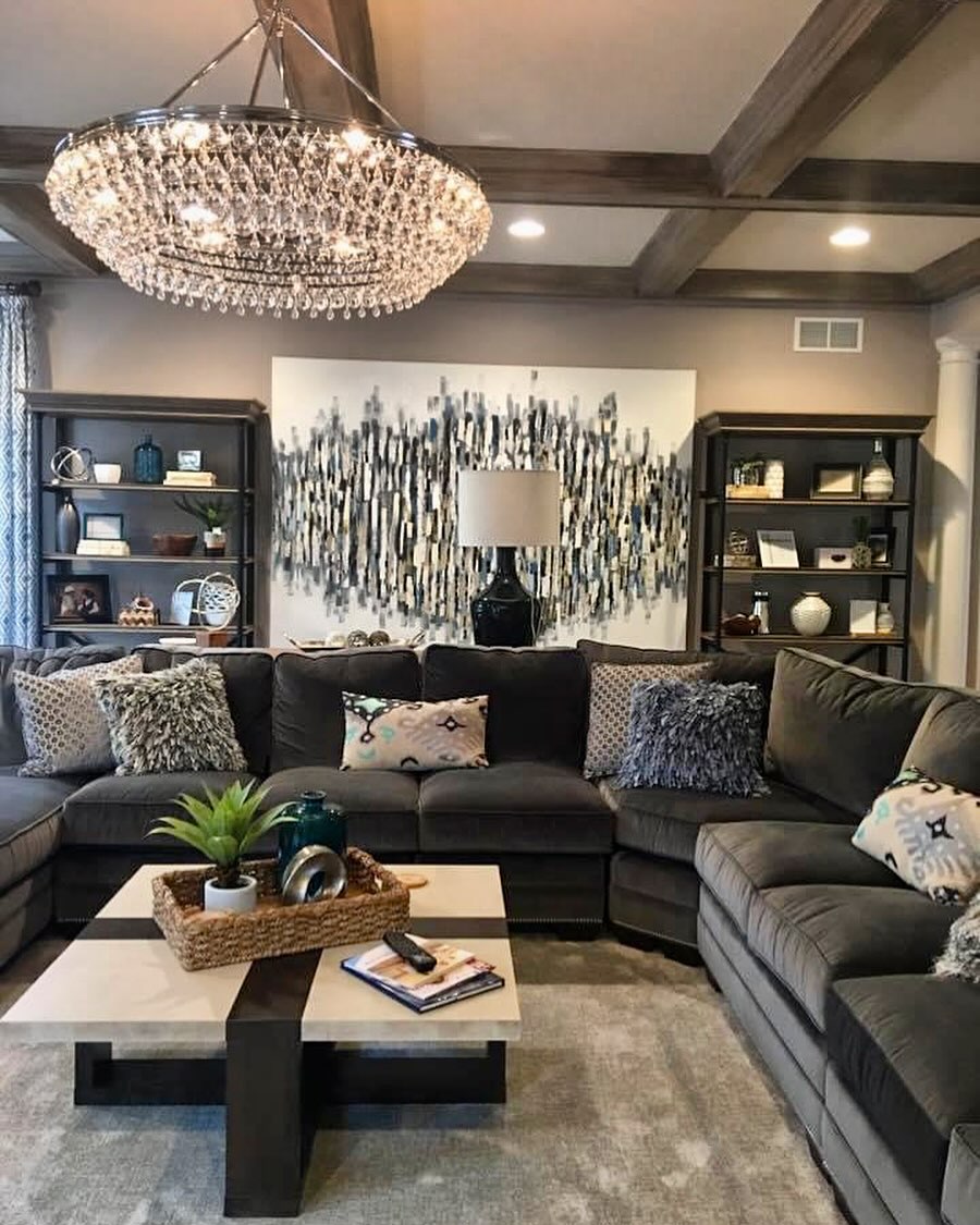 A transitional living room with a dark gray sectional sofa, round crystal chandelier, coffered ceiling with wood beams, abstract blue and gray artwork, two black bookshelves, and a two-tone coffee table.