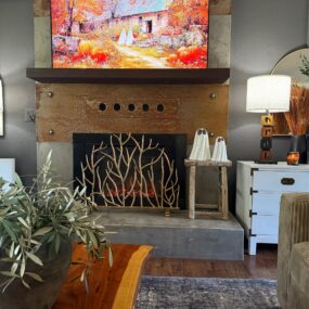 An industrial fireplace with rusted metal surround, mounted TV displaying a fall pumpkin cottage painting, gold branch fireplace screen, two white ghost figurines on a wooden stool, and a live edge coffee table.