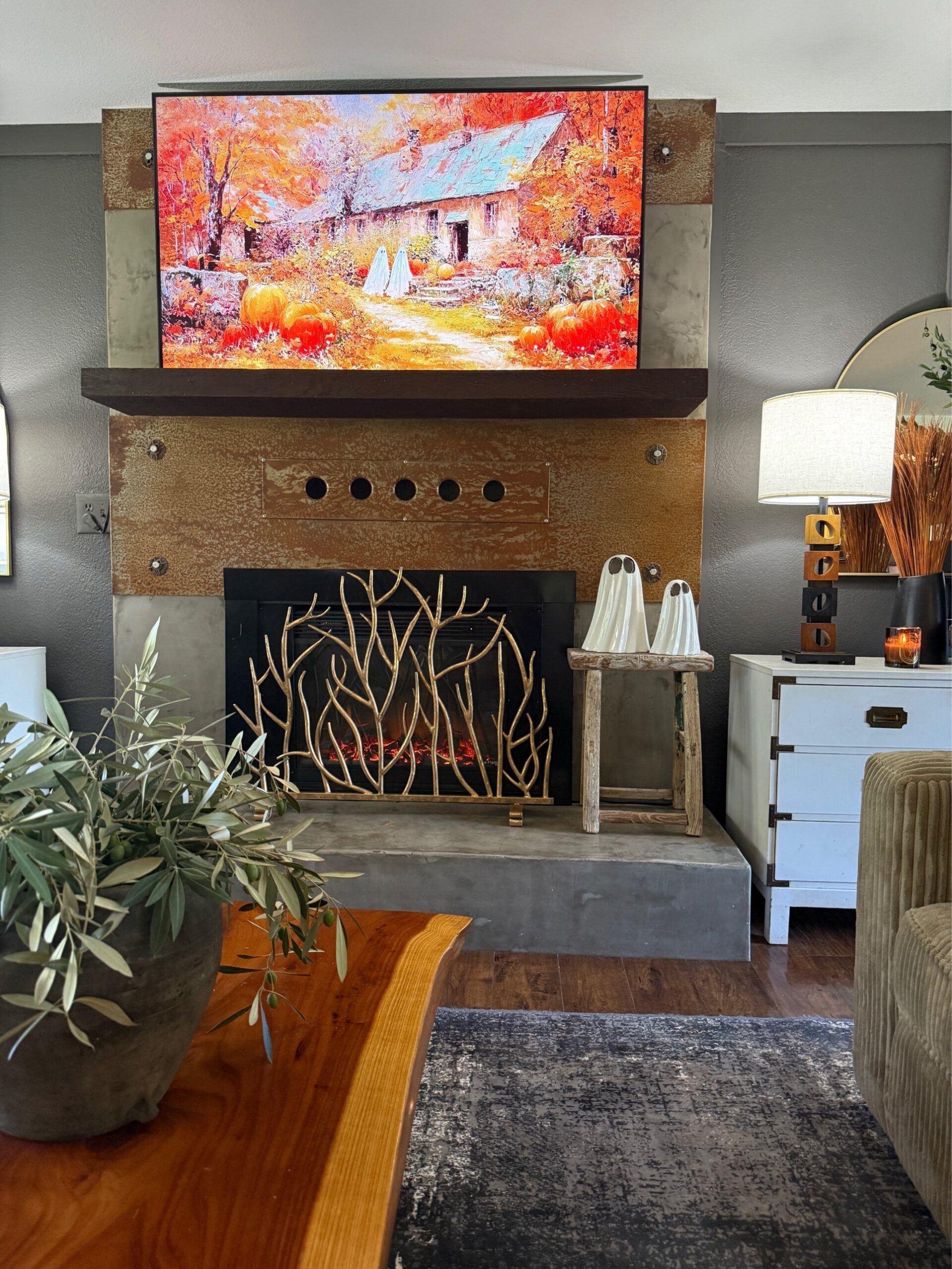 An industrial fireplace with rusted metal surround, mounted TV displaying a fall pumpkin cottage painting, gold branch fireplace screen, two white ghost figurines on a wooden stool, and a live edge coffee table.