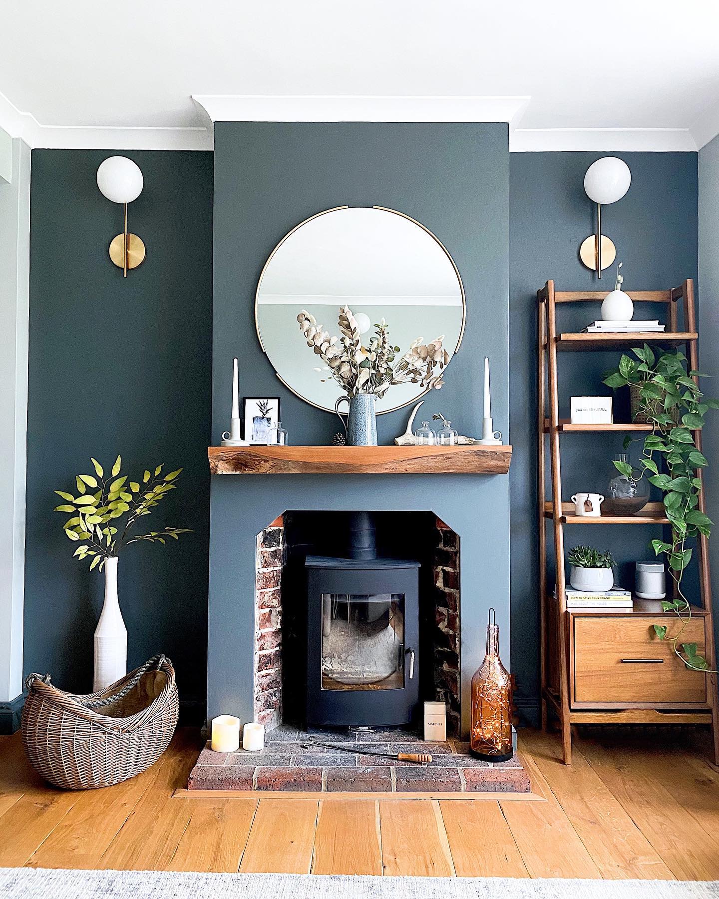 A dark teal living room with a black wood-burning stove in a brick alcove, live edge wood mantel, round gold mirror, globe sconces, wood ladder shelf with trailing plants, and a wicker basket.