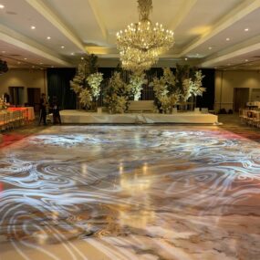 A ballroom with a marble swirl projection-mapped dance floor, large white floral stage installation behind a cream sofa, and a crystal chandelier overhead.