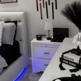 A black and white bedroom with a white tufted bed and blue LED underglow, white nightstand with lamp, and a styled coffee table with black feather plume vase.