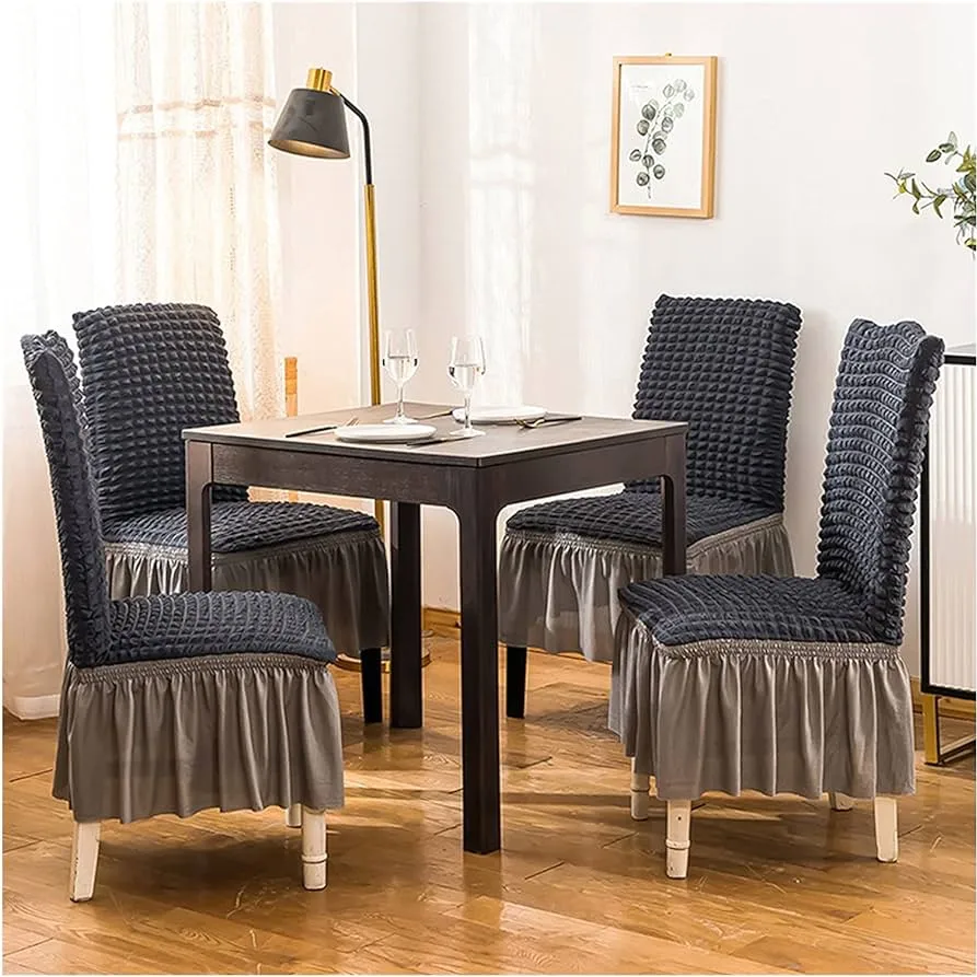 Four dark gray textured dining chair slipcovers with ruffled skirts around a dark wood square dining table set for two.